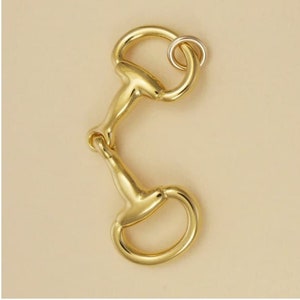 Puede incluir: Un amuleto de bocado de brida de metal dorado con un pequeño anillo adjunto. El amuleto es un accesorio ecuestre popular.
