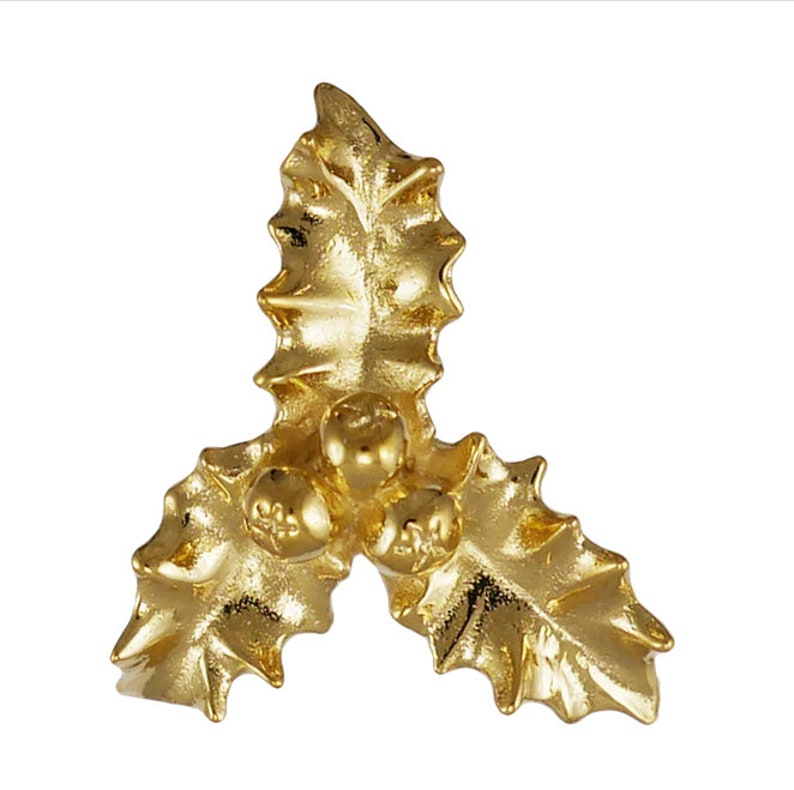 May include: A gold-coloured holly leaf and berry ornament. The ornament features three leaves and three berries, all with a metallic finish. The leaves have detailed veining, and the berries are clustered in the centre.