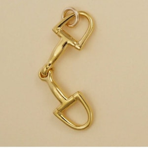 Puede incluir: Un amuleto de metal dorado en forma de bocado de caballo. El amuleto tiene dos bucles, uno más grande que el otro, y un pequeño anillo en la parte superior.