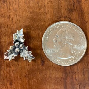 May include: A silver-toned holly leaf and berry charm, next to a U.S. quarter coin. The charm features three berries and detailed leaf textures. The quarter coin is included for size reference.