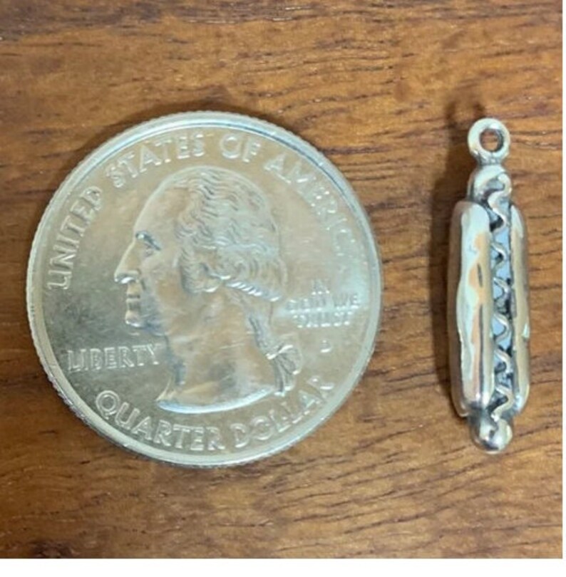 Puede incluir: Un dije plateado con forma de perrito caliente, junto a una moneda de veinticinco centavos de d&oacute;lar estadounidense. El dije tiene un lazo en la parte superior para sujetarlo a una pulsera o collar. El dije de perrito caliente mide aproximadamente 2,5 cm de largo.