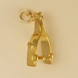 Op de afbeelding: Goudkleurige metalen bedel in de vorm van een cowboy sporen. De bedel heeft een lus aan de bovenkant om aan een armband of ketting te bevestigen.