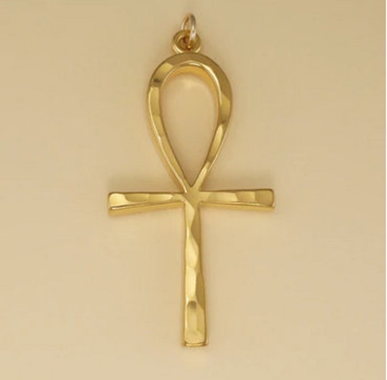 May include: A gold-coloured Ankh pendant with a hammered texture. The Ankh is an ancient Egyptian symbol of life.