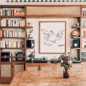 May include: A white dove with colorful flowers drawn in a blue line art style on a white background. The art is framed and hanging on a wall in a room with a wooden bookshelf and a record player. The artist's signature, "Picasso", is written in black below the dove.