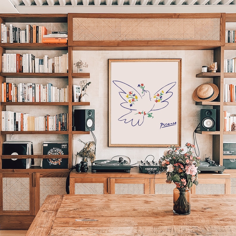 May include: A white dove with colorful flowers drawn in a blue line art style on a white background. The art is framed and hanging on a wall in a room with a wooden bookshelf and a record player. The artist's signature, "Picasso", is written in black below the dove.
