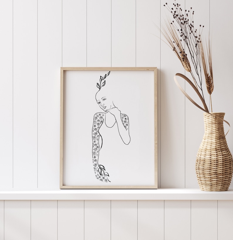 Op de afbeelding: Een zwart-witte lijntekening van een persoon met bloemen op hun armen en een bloemenkroon op hun hoofd. De tekening is ingelijst in een licht houten lijst en hangt aan een witte muur.