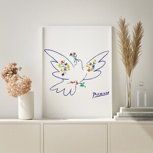May include: A white framed print of a blue line drawing of a dove with colorful flowers. The dove is flying with its wings spread and has a flower in its beak. The artist's signature, "Picasso", is in the bottom right corner.
