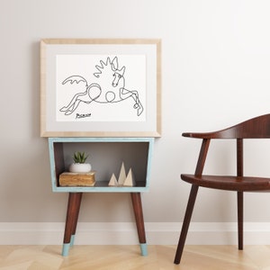 May include: A black and white line drawing of a horse, framed in a light wood frame. The drawing is on a white background and the artist's signature, "Picasso", is visible in the bottom left corner. The frame is hanging on a white wall above a light blue shelf with a small potted plant and wooden decorative pieces.