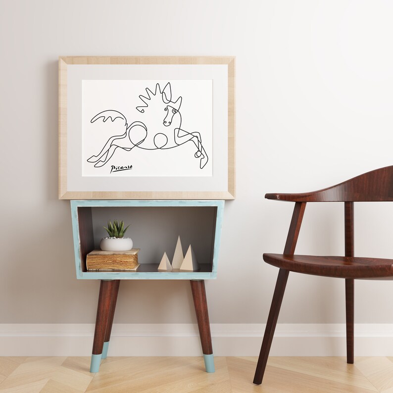 May include: A black and white line drawing of a horse, framed in a light wood frame. The drawing is on a white background and the artist's signature, "Picasso", is visible in the bottom left corner. The frame is hanging on a white wall above a light blue shelf with a small potted plant and wooden decorative pieces.