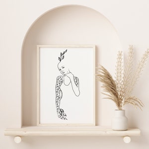 Op de afbeelding: Een zwart-witte lijntekening van een persoon met bloemen die uit hun lichaam groeien. De tekening is ingelijst en hangt aan een muur.