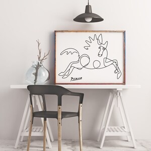 May include: A black and white line drawing of a horse, with the signature "Picasso" below the image. The drawing is framed in a brown wood frame and is hanging on a white wall.