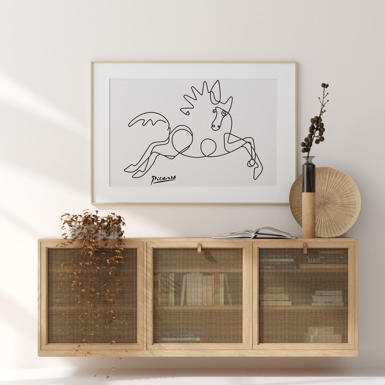 May include: A black and white line drawing of a horse by Pablo Picasso, framed in a white frame and hanging on a white wall above a wooden cabinet with woven wire doors.