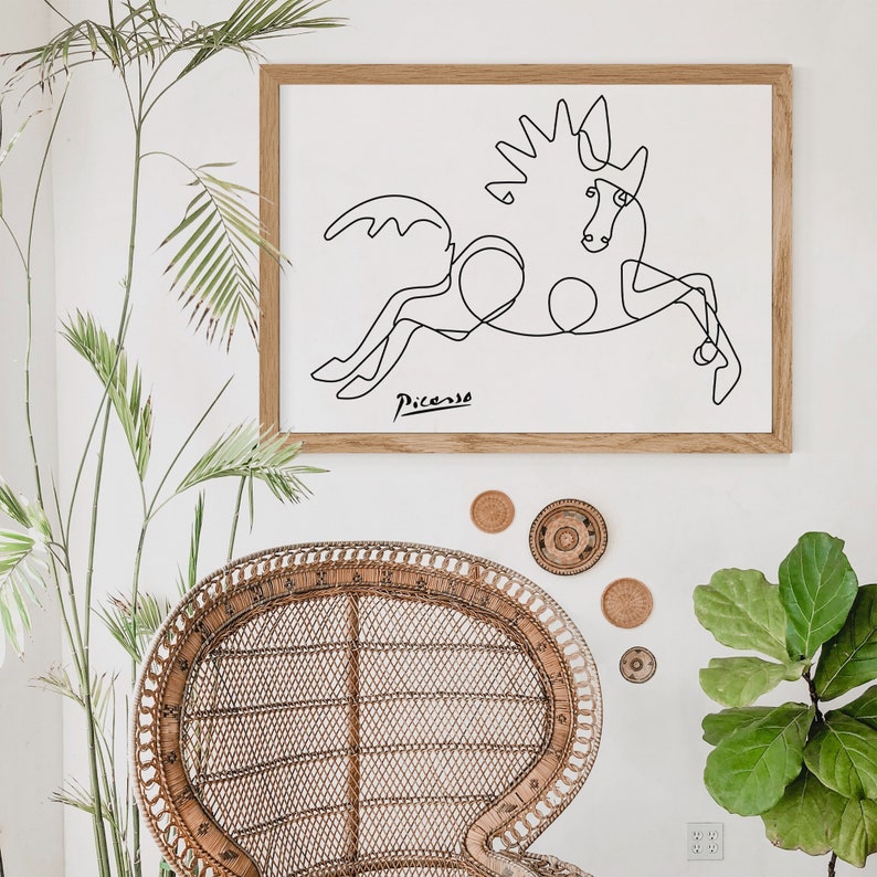 May include: A black and white line drawing of a horse by Pablo Picasso in a wooden frame. The artwork is hanging on a white wall above a wicker chair.