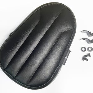 May include: Black motorcycle seat with a quilted design and included mounting hardware. The seat has a textured surface and a stitched border. The mounting hardware includes brackets, washers, and a bolt.
