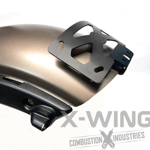 May include: A motorcycle fender in a metallic bronze color with a black metal bracket attached. The image also shows a black component and the text "X-WING COMBUSTION INDUSTRIES".
