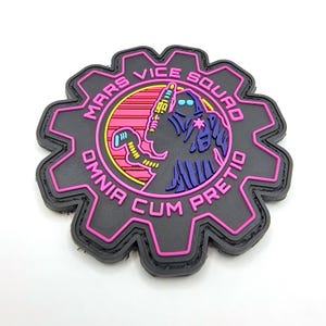 May include: A black and pink patch with a gear design. The patch has the text "Mars Vice Squad Omnia Cum Preto" around the edge. The center of the patch features a cartoon character in a purple and blue suit.