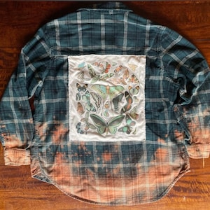 May include: A dark teal plaid button-down shirt with a bleached, faded bottom. The back features a white square with a collage of colorful butterfly illustrations. The shirt has long sleeves and a classic collar.