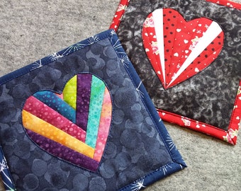 May include: Two handmade quilted heart-shaped pot holders. One has a dark blue background with a rainbow-colored heart. The other has a black and gray background with a red heart. Both have decorative borders.