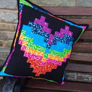 May include: A black cushion with a colourful pixelated heart design. The heart is made up of squares in shades of pink, blue, green, orange, and red. The cushion is sitting on a wooden bench.
