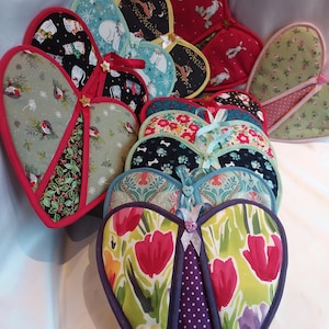 May include: A selection of heart-shaped quilted oven mitts in diverse patterns and colours. Designs include floral, animal, and geometric prints. Each mitt has a contrasting trim and a ribbon or button detail.