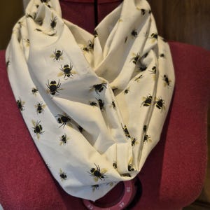 May include: An off-white infinity scarf featuring a repeating pattern of black and yellow bumblebees. The fabric is draped in soft folds, showcasing the detailed insect print across the entire surface of the lightweight material.
