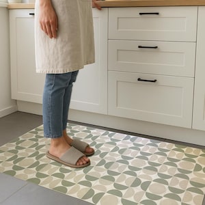 Könnte beinhalten: Ein rechteckiger Küchenteppich mit einem geometrischen Muster in Grün- und Beigetönen. Der Teppich liegt auf einem grauen Boden vor weißen Schränken. Eine Person steht auf dem Teppich und trägt Jeans und Sandalen.