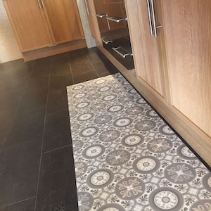 Op de afbeelding: Een gepatroneerd keukenkleed met een geometrisch ontwerp in grijs en wit. Het kleed ligt voor een houten kast met een lade van roestvrij staal.