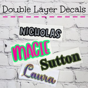 May include: Double Layer Decals are displayed with various names in different fonts and colors. The names include Nicholas, Macie, Sutton, and Laura. The text is layered with contrasting colors, such as black and gray, pink and green, and green and black.
