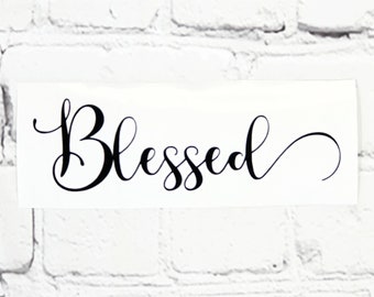May include: A white rectangular sticker with the word "Blessed" in elegant black cursive script. The sticker is against a white brick wall background. The script has a flowing, decorative style.