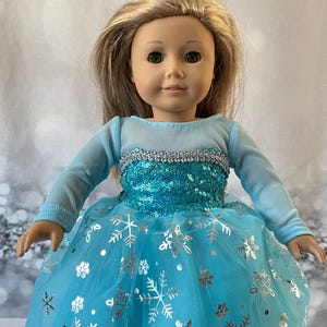 May include: A blue and silver dress with a snowflake pattern for a doll. The dress has long sleeves and a tulle skirt.