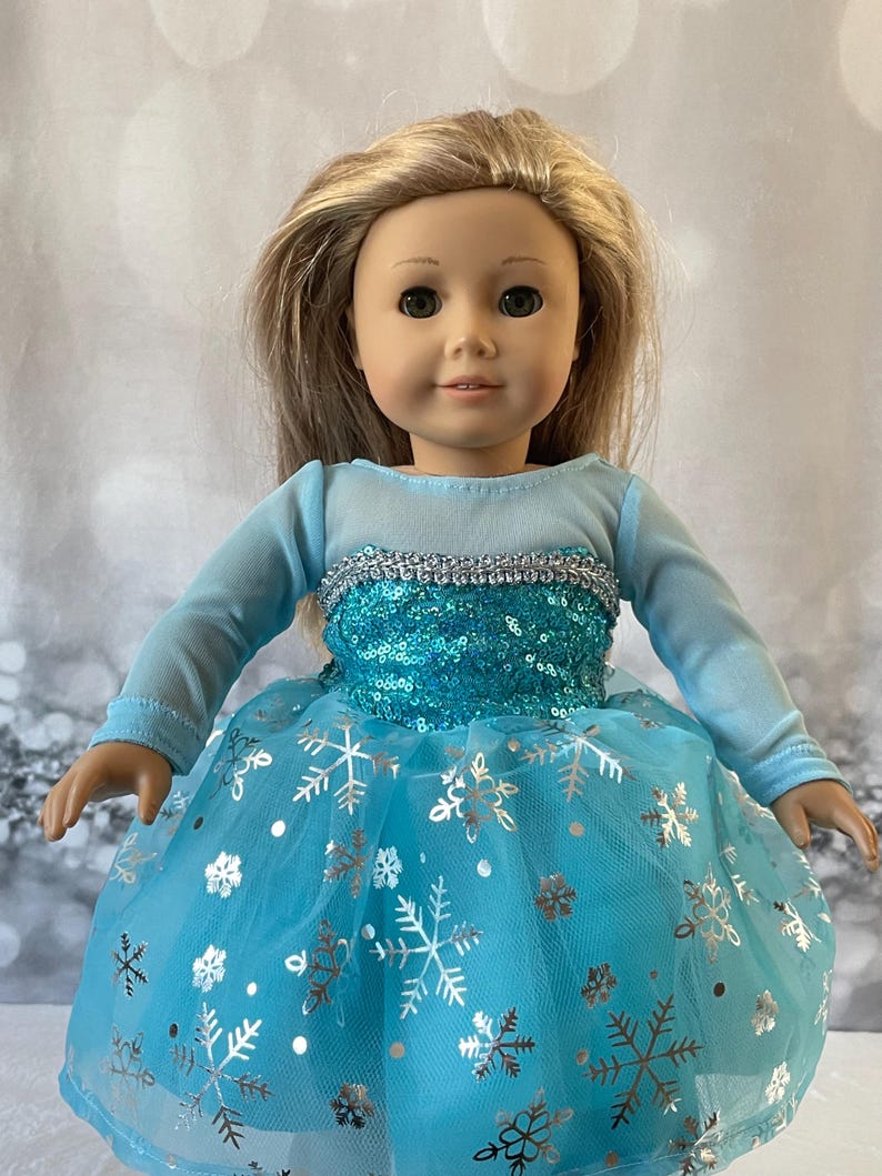 May include: A blue and silver dress with a snowflake pattern for a doll. The dress has long sleeves and a tulle skirt.