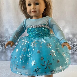 May include: A blue and silver dress with a snowflake pattern for a doll. The dress has long sleeves and a tulle skirt.