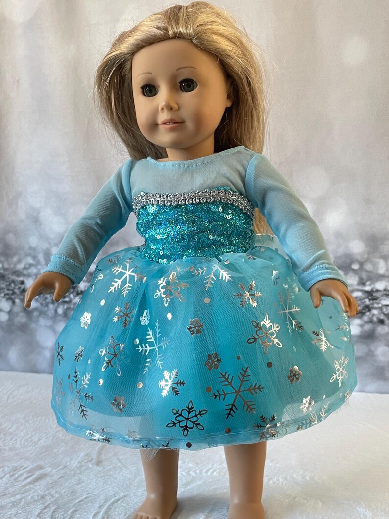 May include: A blue and silver dress with a snowflake pattern for a doll. The dress has long sleeves and a tulle skirt.