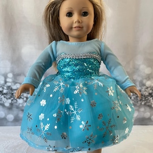 May include: A blue and silver snowflake dress for a doll. The dress has a tulle skirt with silver snowflakes and sequins on the bodice.