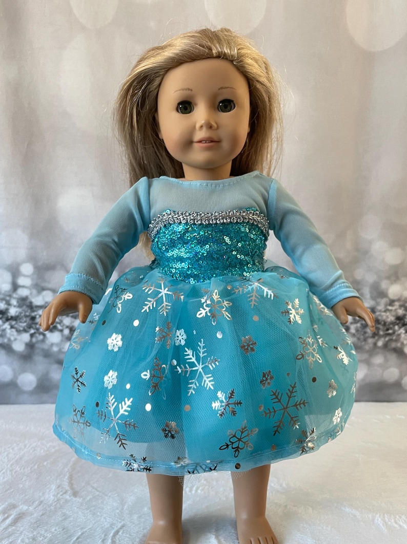 May include: A blue and silver snowflake dress for a doll. The dress has a tulle skirt with silver snowflakes and sequins on the bodice.