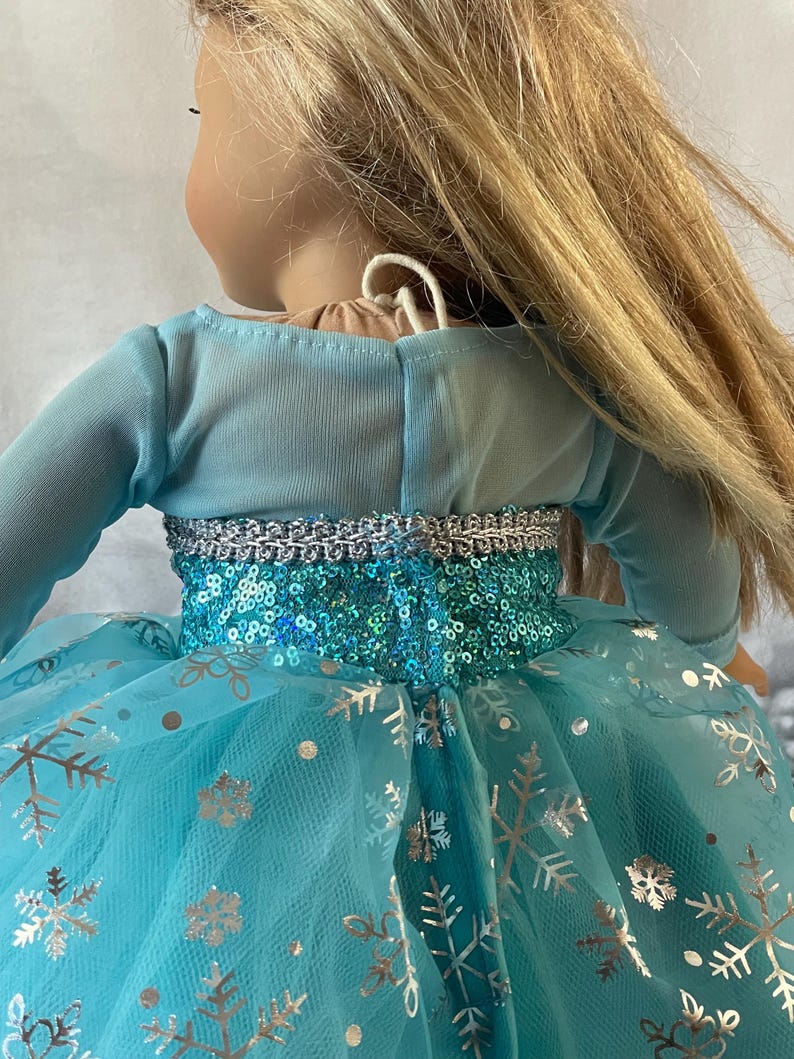 May include: A blue tulle dress with silver snowflake print and a sequined bodice. The dress has long sleeves and a fitted bodice.
