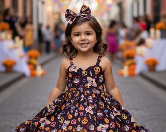 May include: A young girl smiles, wearing a dark purple dress with a colorful Day of the Dead skull print. The dress has a ruffled hem and spaghetti straps. A matching bow adorns her hair. The background features a festive street scene with colorful decorations.