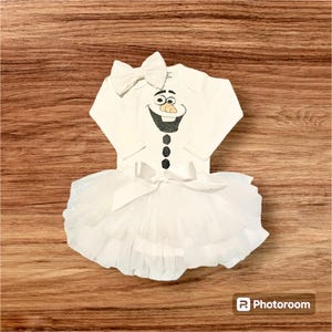 May include: A white baby outfit featuring a long-sleeved onesie with a cartoon snowman face and a white tutu skirt. The snowman face has black glitter accents and a carrot nose. A white bow is attached to the onesie and the skirt.
