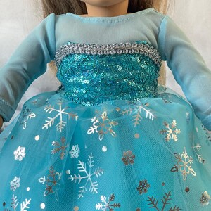 May include: A blue tulle dress with silver snowflake print and a sequined bodice. The dress has long sleeves and a silver trim around the neckline.