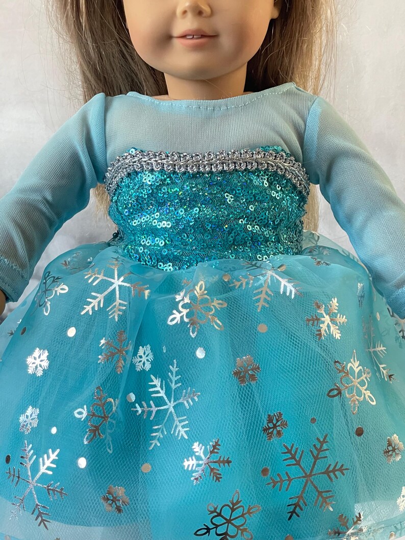 May include: A blue tulle dress with silver snowflake print and a sequined bodice. The dress has long sleeves and a silver trim around the neckline.