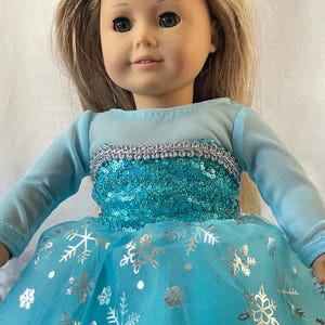 May include: A doll wearing a blue and silver dress with a sequined bodice and a tulle skirt with snowflake designs.