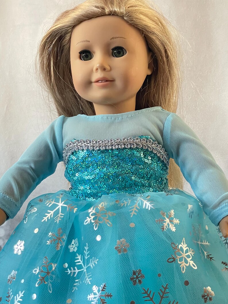 May include: A doll wearing a blue and silver dress with a sequined bodice and a tulle skirt with snowflake designs.
