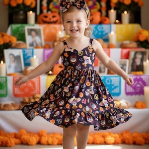 May include: A young girl wearing a dark blue dress with a colorful Day of the Dead print, including skulls and flowers. She also wears a matching headband. The dress has a ruffled hem and spaghetti straps. She is smiling and standing in front of a decorated altar.