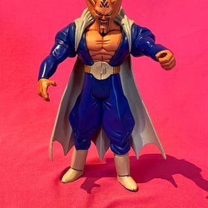 May include: A detailed action figure of a character with orange skin, a blue outfit, and a white cape. The figure has a stern expression and is posed with arms outstretched. The figure is standing on a pink surface.