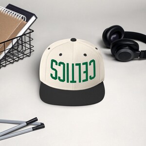 May include: A cream-colored baseball cap with a black brim. The word "CELTICS" is printed in green block letters across the front. A pair of black headphones and a wire basket with notebooks are in the background.