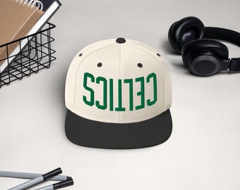 Può includere: Un cappellino da baseball color crema con visiera nera. La parola "CELTICS" è stampata in lettere verdi sul davanti. Un paio di cuffie nere e un cestino di filo metallico con quaderni sono sullo sfondo.