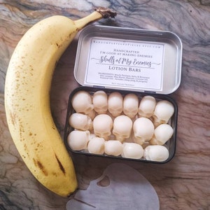 May include: A banana next to a tin containing skull-shaped lotion bars. The tin has a label that reads "Skulls of My Enemies Lotion Bars." A paper skull stencil is also present. The banana is approximately 7 inches long.