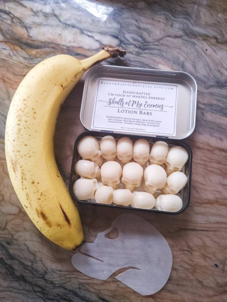 May include: A banana next to a tin containing skull-shaped lotion bars. The tin has a label that reads "Skulls of My Enemies Lotion Bars." A paper skull stencil is also present. The banana is approximately 7 inches long.