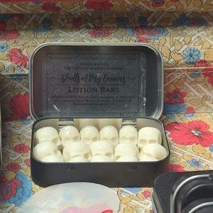 May include: A black metal tin filled with skull-shaped lotion bars. The tin's label reads "Skulls of My Enemies LOTION BARS." The lotion bars are white and arranged neatly inside the open tin. The background features a floral pattern.