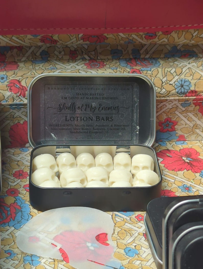 May include: A black metal tin filled with skull-shaped lotion bars. The tin's label reads "Skulls of My Enemies LOTION BARS." The lotion bars are white and arranged neatly inside the open tin. The background features a floral pattern.